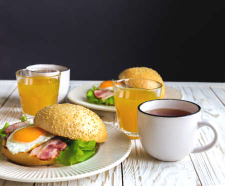 Burger sandwich with sesame bun, orange yolk fried egg, bacon and green salad, cup of tea and orange...