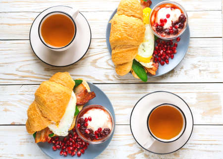 Fresh croissant  with orange yolk fried egg, bacon, spinach and cheese, homemade yogurt with cherry syrop, pomegranate and cup of tea for breakfast. Selective focus, top view. Continental breakfast conceptの写真素材