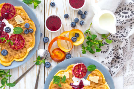 Cup of coffee and Belgium waffles with blueberry, blood orange,  whipped cream, jam, mint and white chocolate on wooden table. Breakfast concept.の写真素材