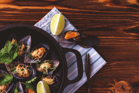 Seafood. Baked shellfish mussels with cheese, cilantro and lime in half shells in cast iron black pan on dark wooden background. Healthy food concept. Top view.の写真素材