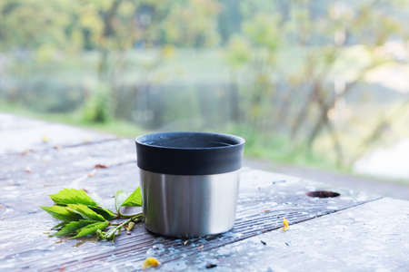 Thermal cup of coffee on wooden table with nature on background.