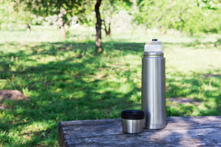 Thermal bottle and cup of coffee on wooden table with nature on background.