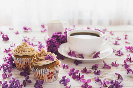 Homemade tiramisu cupcakes with whipped cream, cup of coffee, notebook  and bouquet of purple lilac spring flowers on rustic wooden backgroundの写真素材