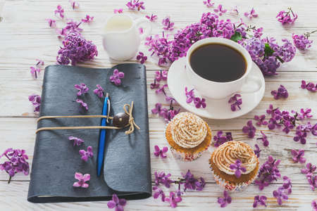 Homemade tiramisu cupcakes with whipped cream, cup of coffee, notebook  and bouquet of purple lilac spring flowers on rustic wooden backgroundの写真素材