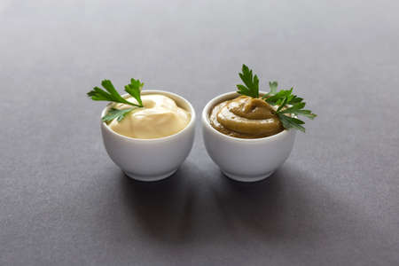 Fresh homemade sauces yellow mustard and white mayonnaise with spices and parsley on dark...