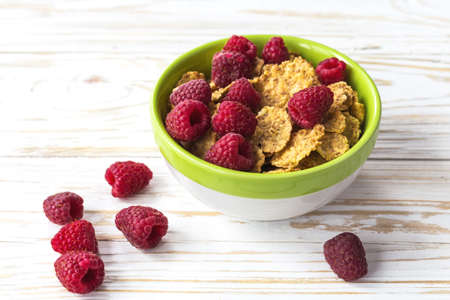 Bowl with cereals, raspberries and blueberries. Healthy corn flakes breakfast on the white wooden...