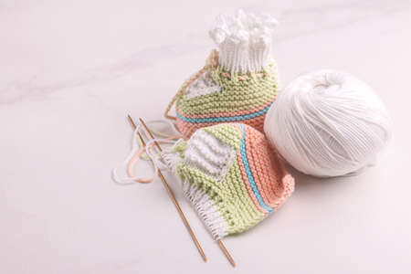 Knitted baby booties with yarn and needles on white marble background. Warm handmade socks for the...