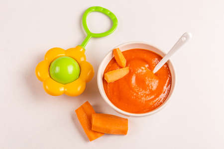 Pureed carrot baby food in clay bowl with raw carrot and beanbag on white marble background. Copy spaceの写真素材