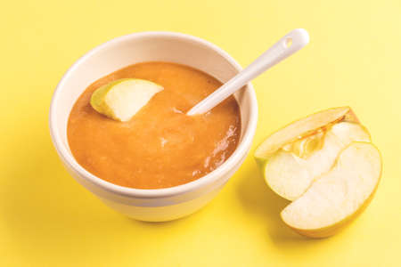 Pureed apple baby food in clay bowl with raw apple and beanbag on bright yellow background. Copy...