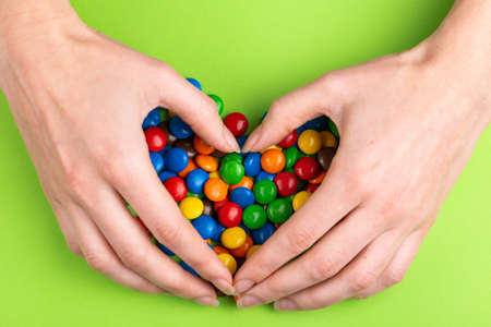 Glazed colored chocolate colored candies are gathered into heart by woman's hands on bright green...
