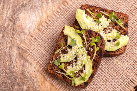 Healthy rye toasts with avocado, radish sprouts and sesame on burlap napkin. Wooden background....