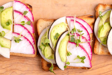 Tasty wheat toasts with radish, avocado and sprouts on wooden cutting board. Dark concrete background. Simple healthy breakfast. Copy space. Top viewの写真素材