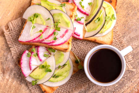 Delicious wheat toasts with radish, avocado and sprouts, cup of strong coffee on burlap napkin. Wooden background. Simple healthy breakfast. Top viewの写真素材