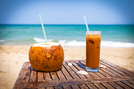 Coconut drink and ice coffee on the wooden table against beautiful beach and emerald seaの写真素材