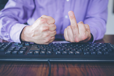 Agression in the Internet, man sitting behind keyboard with hand clenched into a fist and middle finger extendedの写真素材