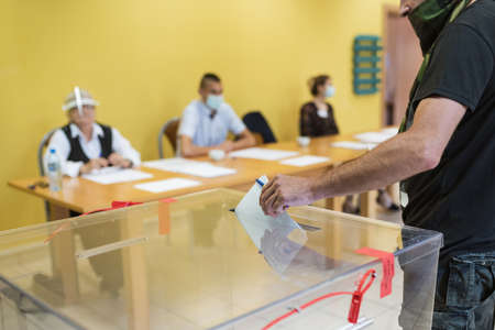Throwing a card with a vote
to the ballot box during  elections. In the backround election commission with face mask due to pandemic Covid-19.の写真素材