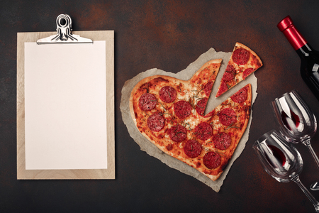 Heart shaped pizza with mozzarella, sausagered, wine bottle, two wineglass and tablet on rusty background. Top viewの写真素材