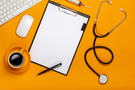 Top view of a doctor's table with notepad and pen stethoscope, keyboard, prescription and pills, a cup of coffee on a yellow background with space for your text.の写真素材