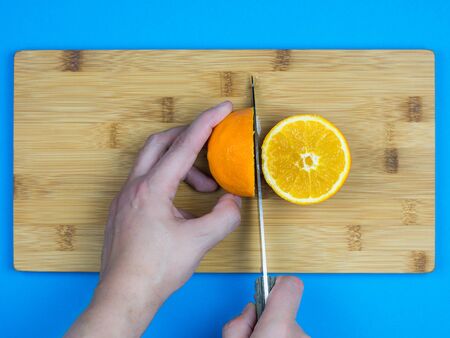 caucasian man using a knife to cut an orange on the top of a wooden surface with blue backgroundの写真素材