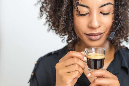 transparent cup with black creamy espresso coffee hold by a beautiful afro black woman with a helpdesk headset. selective focus on cupの写真素材