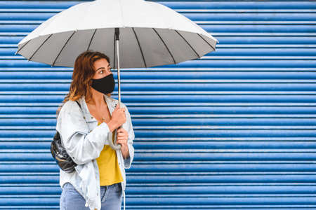 Caucasian young woman standing near blue grid background with an umbrella and black face maskの写真素材