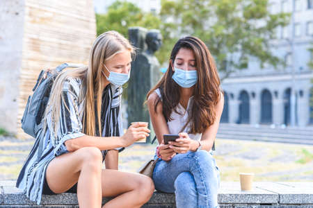 two young beautiful female friends sitting on a stone bench in the street and wearing blue hygienic masks showing each other smart phone screenの写真素材