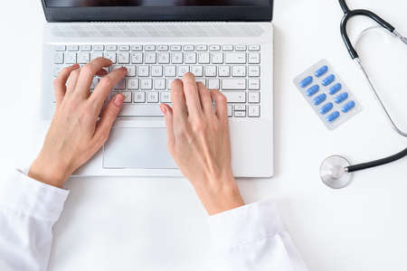 top view of the hands of a caucasian doctor using a modern laptop on the top of a white desk