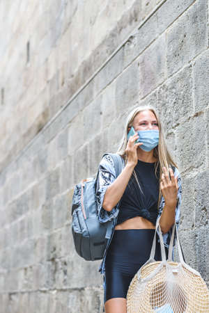 young beautiful caucasian woman wearing a face mask talking by phone in street near a wall with big copy space on the topの写真素材