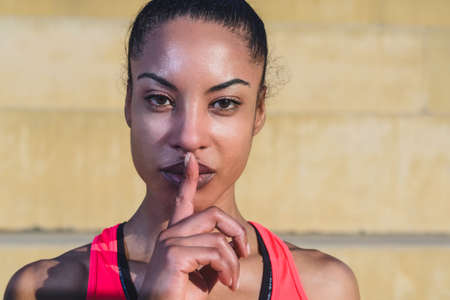 portrait of attractive young African woman wearing sportswear looking at camera and making silence gesture with a finger on her lips. copy space on the rightの写真素材