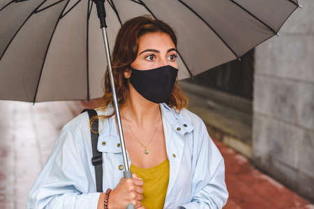 caucasian redhead young woman with casual clothes and wearing a black protective mask holding an umbrella in the streetの写真素材