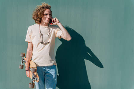 young attractive skater taking with his smart phone near a green wall on a sunny day