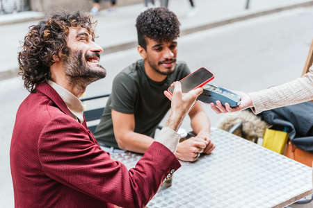 Attractive caucasian man sitting with latin friend and paying bar bill with his smart phoneの写真素材