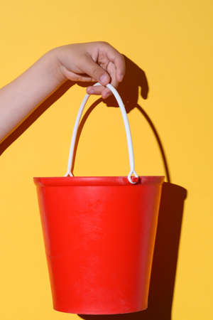 red toy bucket on the hands of a caucasian child with yellow backgroundの写真素材