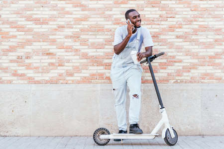 portrait of an african young man with a scooter talking by phone in the street