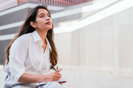 thoughtful indian business woman sitting with a notebook and a pen on a bench outdoorsの写真素材