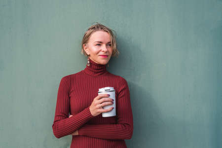 caucasian blonde woman taking a break with a coffee near a industrial green wall
