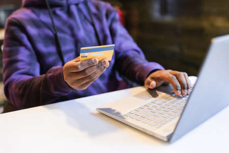 Crop anonymous African American male typing bank card data for online paying for online purchases in...