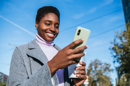Black woman using mobile phone against blue skyの写真素材