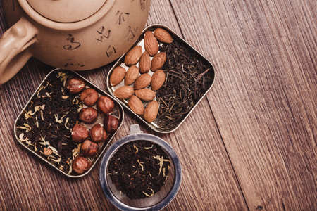 The composition of the tea on a wooden surface. Eco-friendly brown teapot from clay with a scattering of tea, almond and hazelnut. Green and black tea in metal boxes. Top view with copy spaceの写真素材