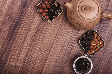 The composition of the tea on a wooden surface. Eco-friendly brown teapot from clay with a scattering of tea, almond and hazelnut. Green and black tea in metal boxes. Top view with copy spaceの写真素材