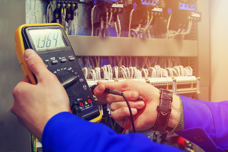 Multimeter in hands of electrician close-up against  background of electrical wires and relays. Adjustment of scheme of automation and control of electrical equipmentの写真素材