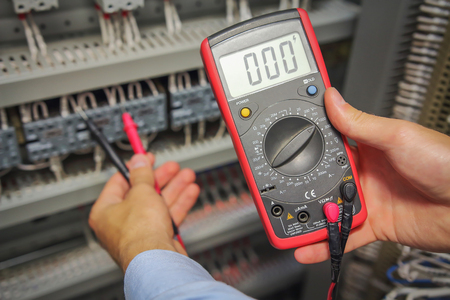 Multimeter in hands of electrician close-up on blurred background of electrical elements. engineer performs adjustment work in control cabinet of industrial equipment.の写真素材