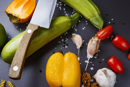 Vegetables on black background for cooking delicious dish. Kitchen knife and raw vegetables pepper, zucchini, garlic, lime, walnuts, spices, tomatoes. Components of healthy dietの写真素材