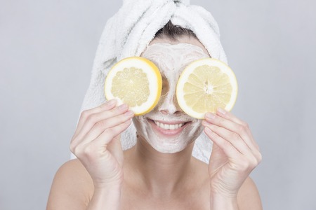 Smiling brunette woman holding two slice of lemon in front of her face. woman with moisturizing facial mask. Beauty and skin care conceptの写真素材