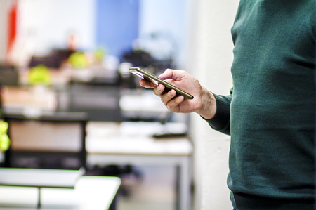 The office employee uses a modern smartphone in the background of the office. The employee of the corporation with a mobile phone in the hands on the background of the workspaceの写真素材