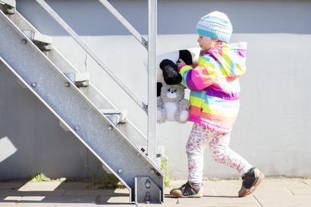 little girl is playing with plush toys near metal staircase on bright sunny afternoon.