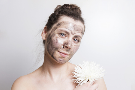 Portrait of attractive brunette girl with black cosmetic face mask and holds white flower. Facial mask skin care. SPA treatmentsの写真素材