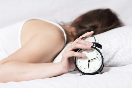 girl is sleeping on bed in bedroom and turns off alarm clock. woman does not want to wake up in early morning. girl turned away from alarm clock. Early rise to work. Female sleepの写真素材