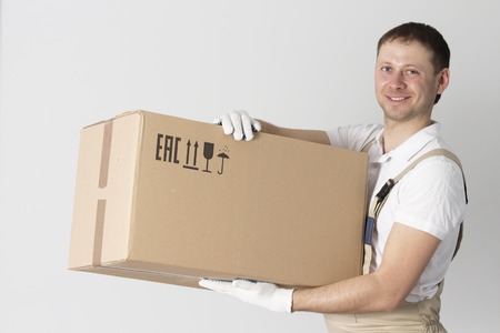 Portrait of mover with cardboard box in hands on gray background. Relocation services man holds box. Loader in uniformの写真素材