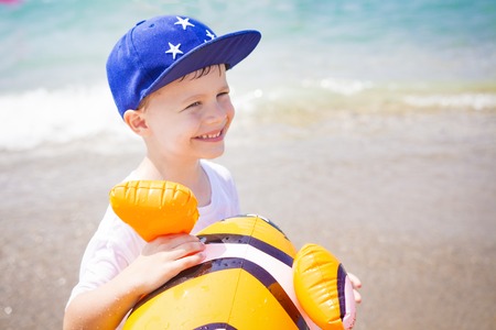 Portrait of smiling happy boy in hat with toy fish balloon on blue sea beach background. Happy boy on sea. Child on tropical vacation.の写真素材